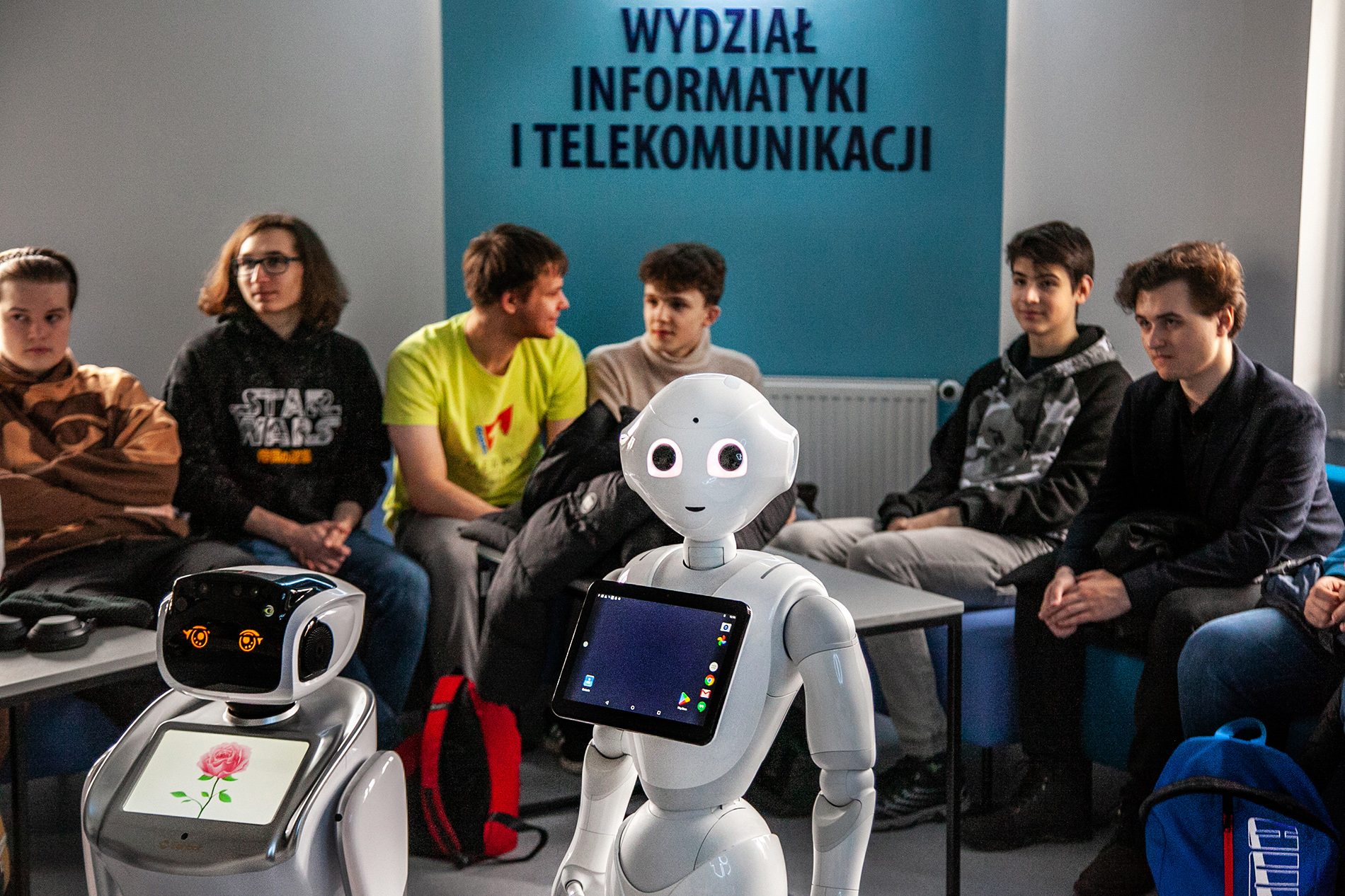 2 roboty humanoidalne w sali Wydziału Informatyki i Telekomunikacji, a w tle uczniowie